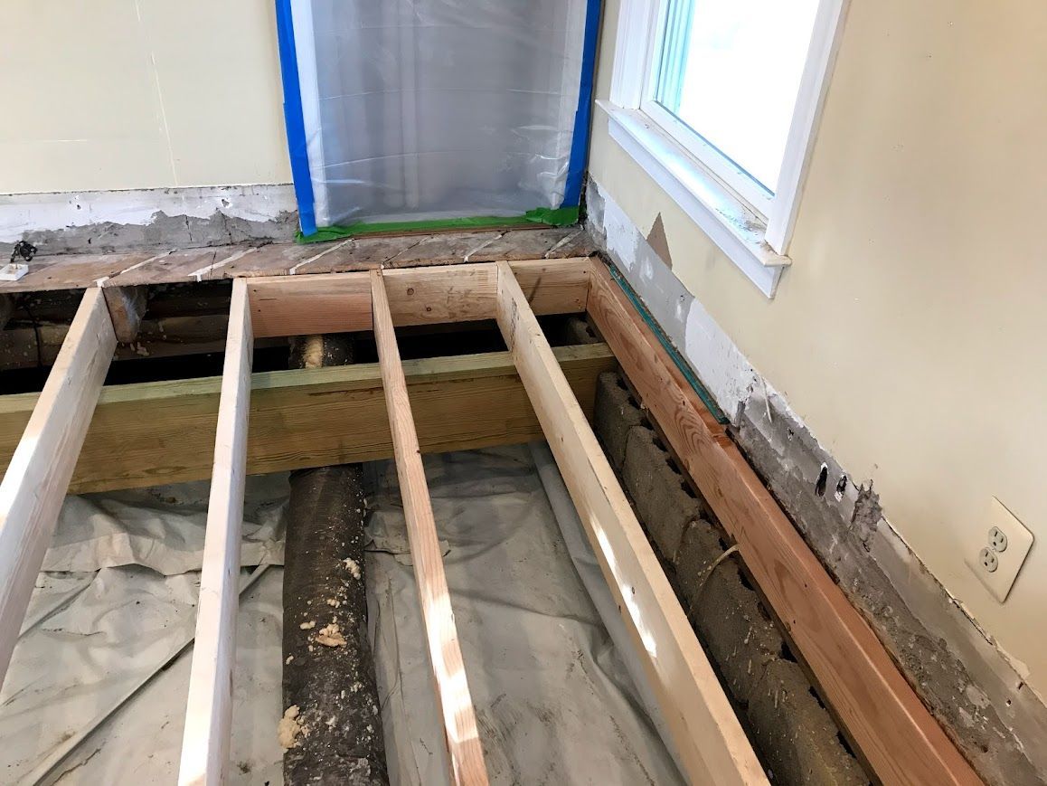 Interior view showing floor joists exposed during a renovation. White and tan wooden beams, insulation. Wall with window.