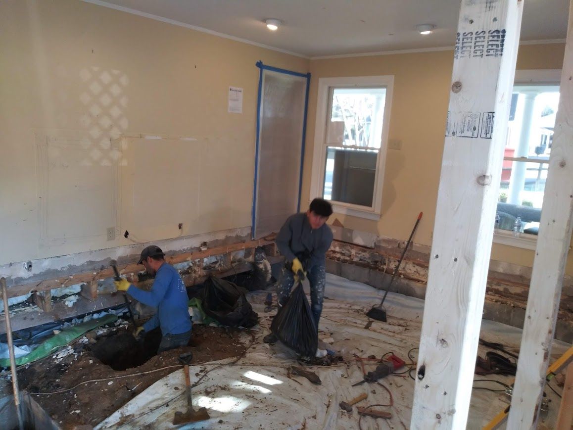 Two people in a room under renovation, removing debris from the floor, with exposed studs and unfinished walls.