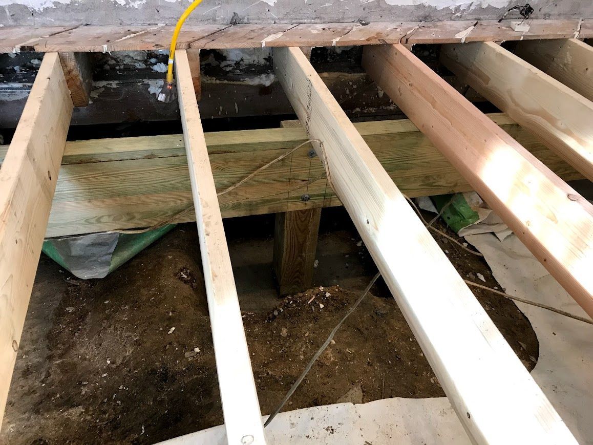 Wooden floor joists exposed in a construction site, with dirt and debris visible below.