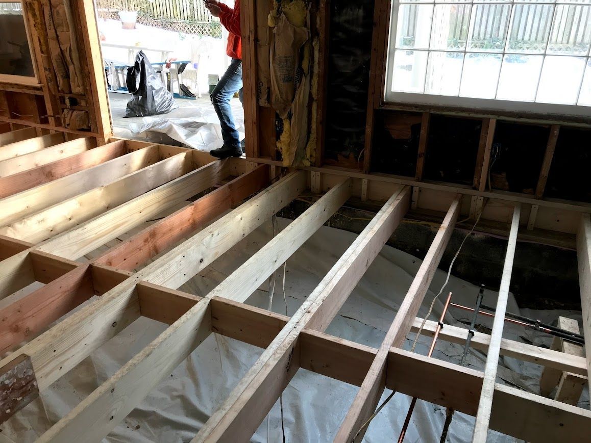 Interior view of a room under construction, showing exposed floor joists. A person is standing near a window.