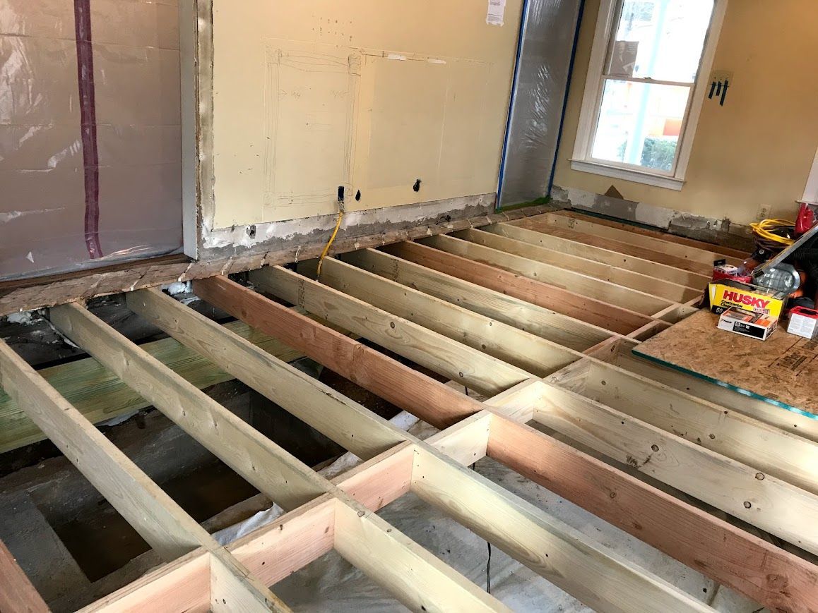 Wooden floor joists exposed in a room during construction. Wall with insulation visible. Window in the background.