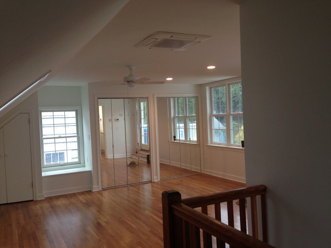 Spacious room with hardwood floors, windows, and mirrored closet. Features a ceiling fan and white walls.