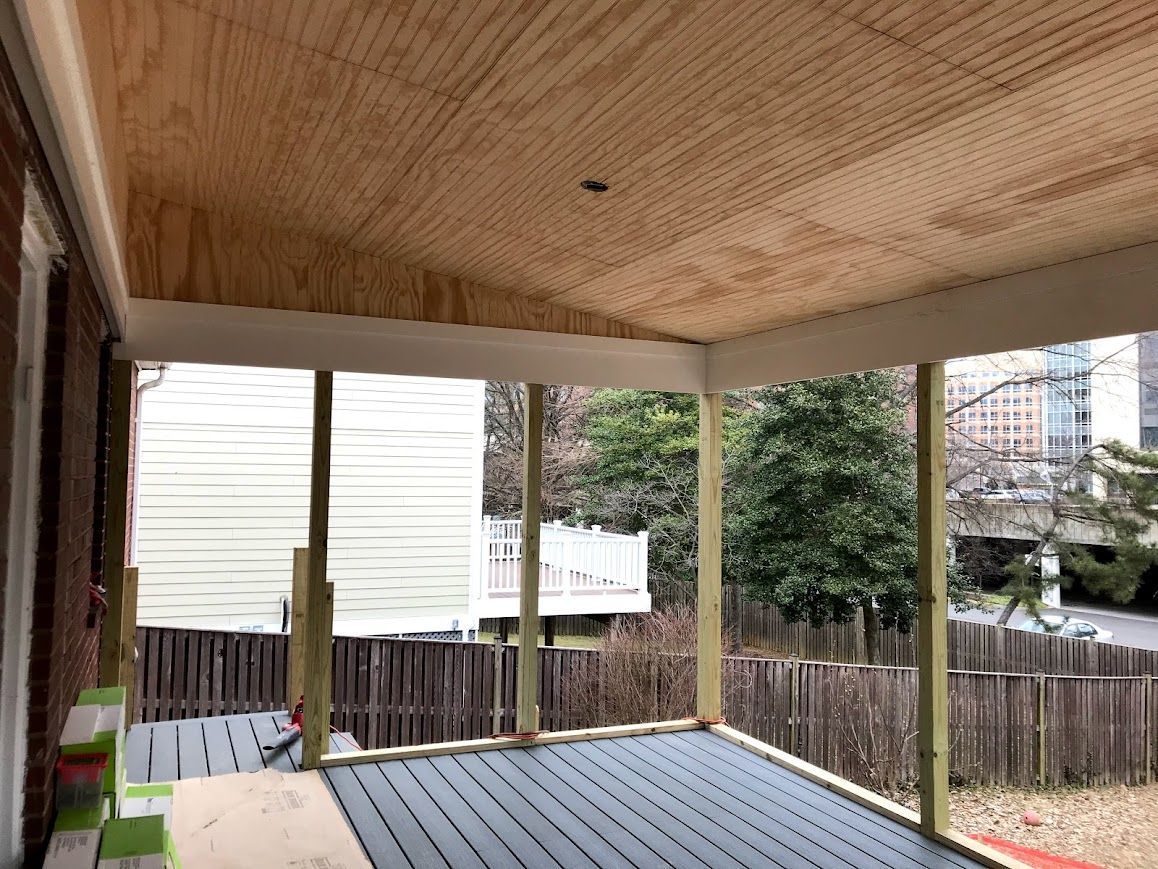 Porch with wooden ceiling, support beams, and a gray-slatted floor, overlooking a backyard and buildings.