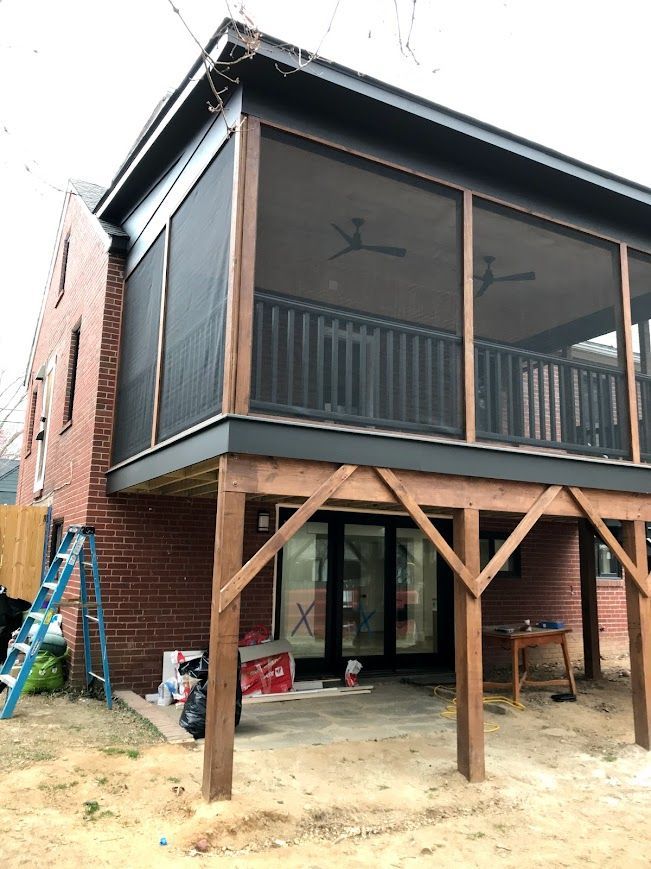 Screened porch with dark framing and railings, attached to a brick house. Supported by wooden beams.