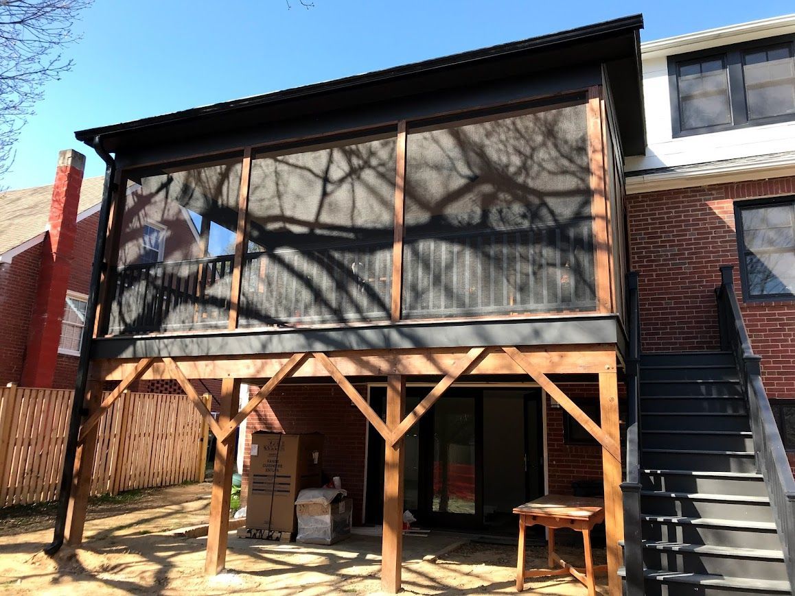 Screened-in porch on elevated wooden deck, attached to a brick building. Black roof, brown trim, and wooden stairs.