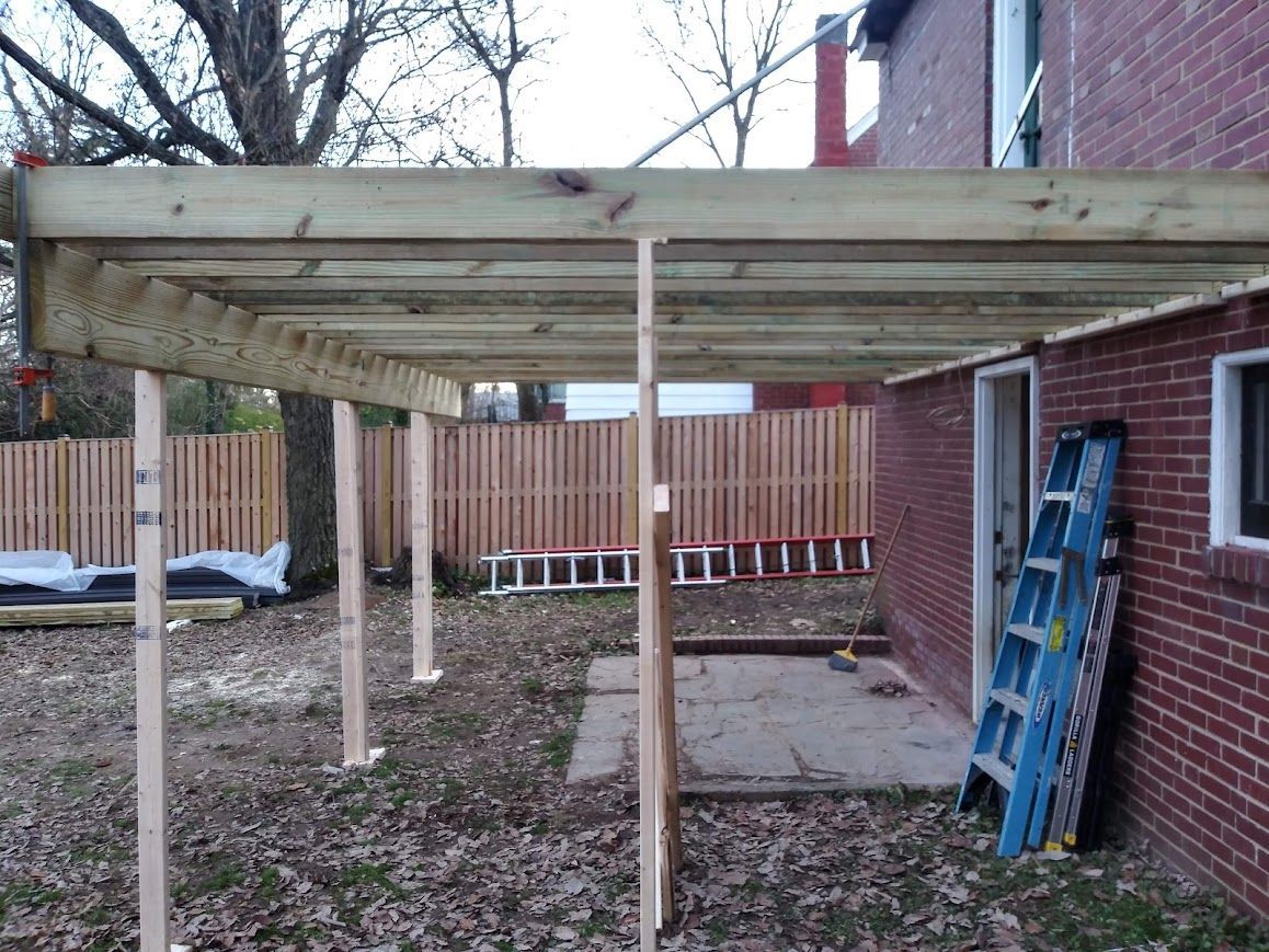 Construction of a covered patio with wooden beams and support posts next to a brick building.