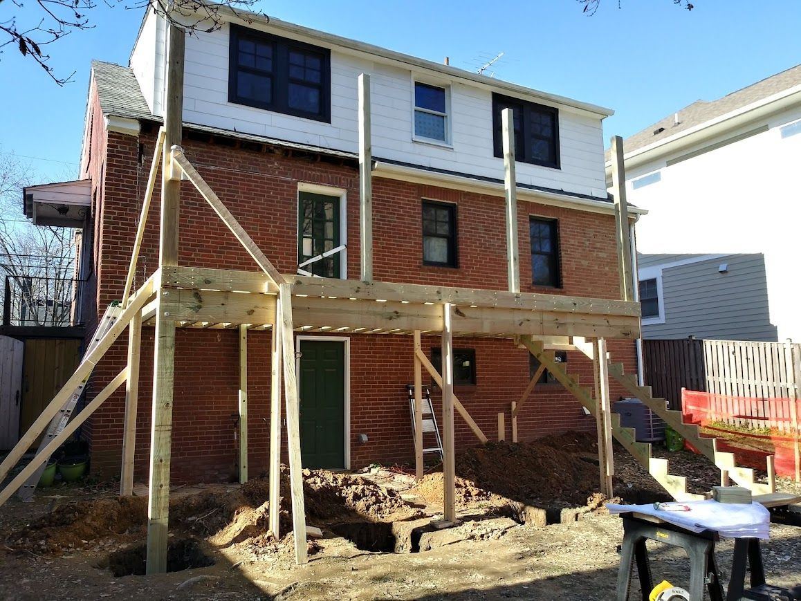 Deck construction: wooden deck frame attached to a brick building; ground dug out below.
