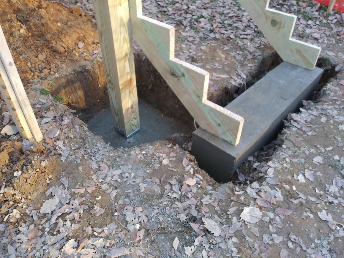 A wooden staircase under construction, set in dirt. Concrete supports the base and post.