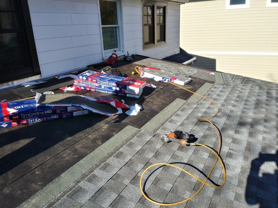 Roof with shingles being replaced; bundles of new shingles and a nail gun are present.