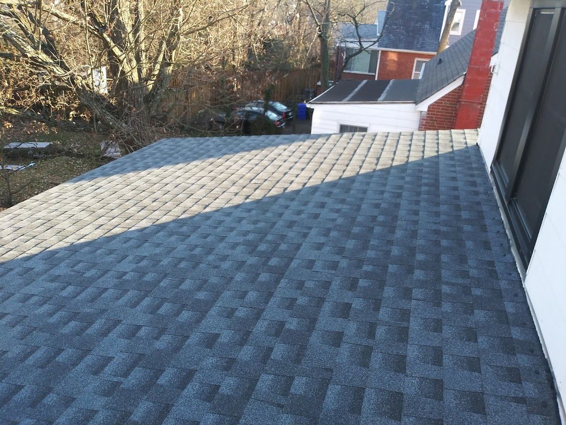 View of a newly shingled roof with a dark gray color, trees, and parts of neighboring houses in the background.