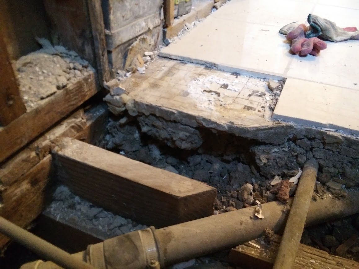Damaged floor, exposed pipes, and debris. Tiles lifted, revealing underlying structure in corner.