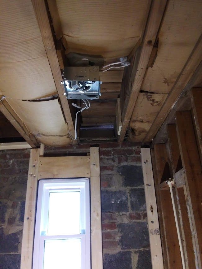 Ceiling with exposed joists, light fixture, damaged plywood, brick wall, and window frame.