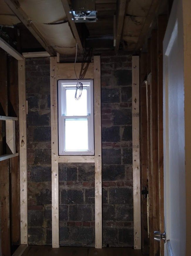 Interior view of a room under construction with a window centered on a brick wall, wood framing visible.
