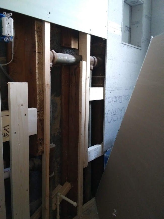 Interior view of exposed plumbing pipes within a wall. Wooden framing and electrical outlets are visible.
