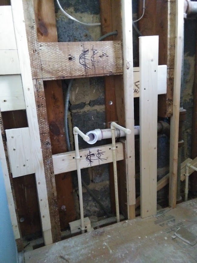 Bathroom wall with exposed plumbing, white pipes, and wooden framing.