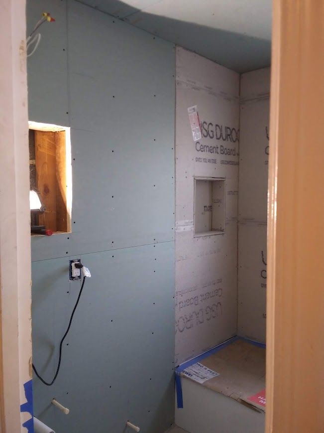 Bathroom under construction, cement board walls with a built-in shelf, blue tape, and a small window.