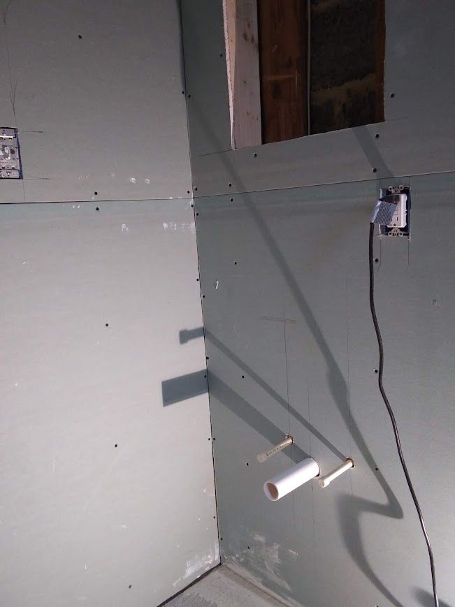 Corner of a room with unfinished drywall. Electrical outlet and exposed framing. Shadow of object on wall.