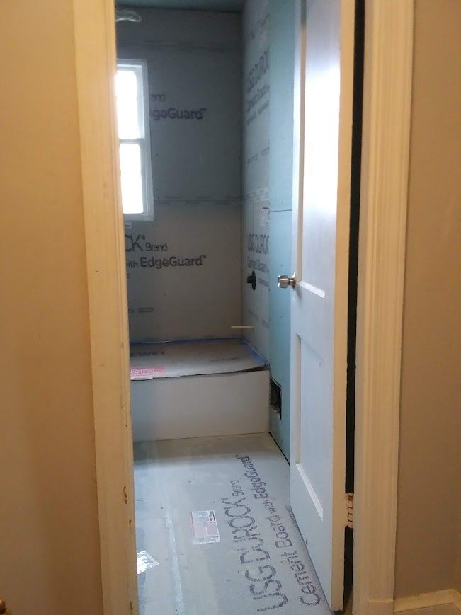 Bathroom under construction with shower area featuring blue wallboards, a window, and a door on the right.