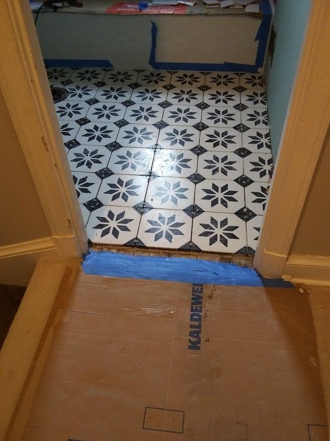 Black and white floral tile floor in a doorway, with blue painter's tape and cardboard protection.