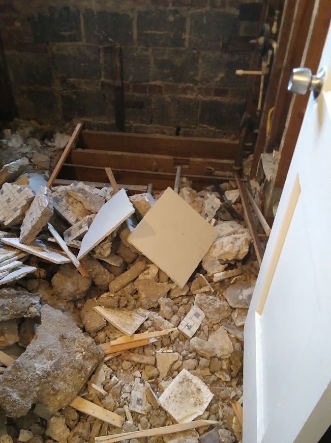 Demolished bathroom interior; debris on floor, exposed brick wall, door frame visible.
