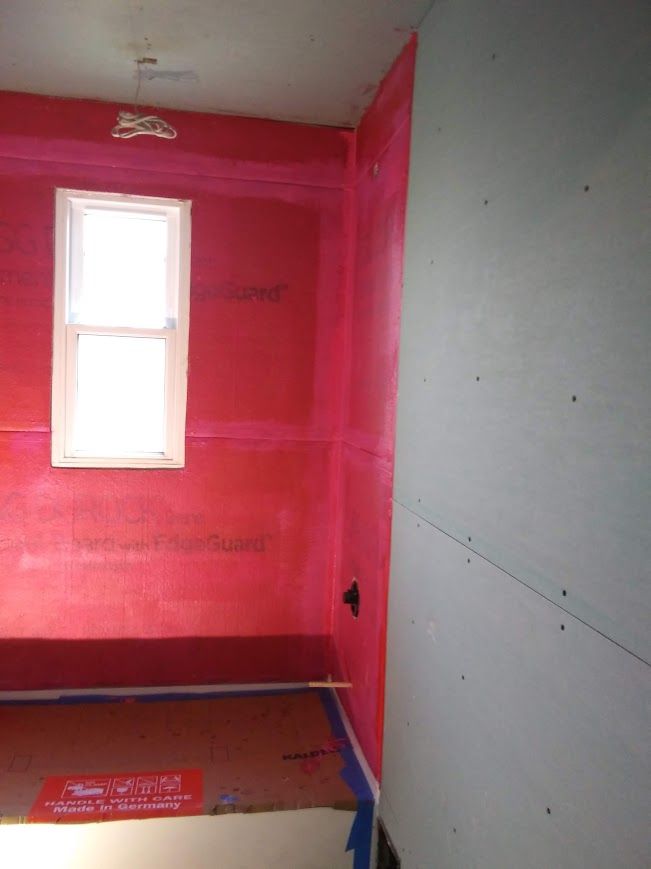 A bathroom with red waterproofing on the walls and floor, a white window, and a gray wall on the right.