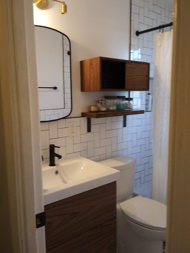 Small modern bathroom with white tile, wooden vanity, and floating shelf.