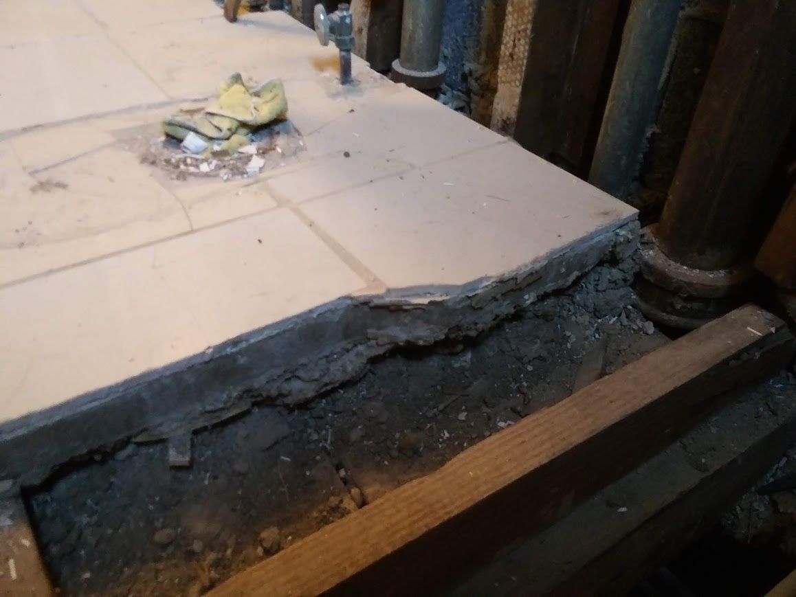 Damaged tile floor edge, revealing concrete and wooden support beams in a room.