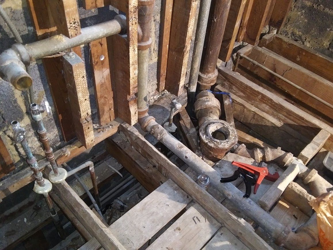 Exposed plumbing pipes in a wall cavity under construction. Rusty pipes, wood framing, and a red tool visible.