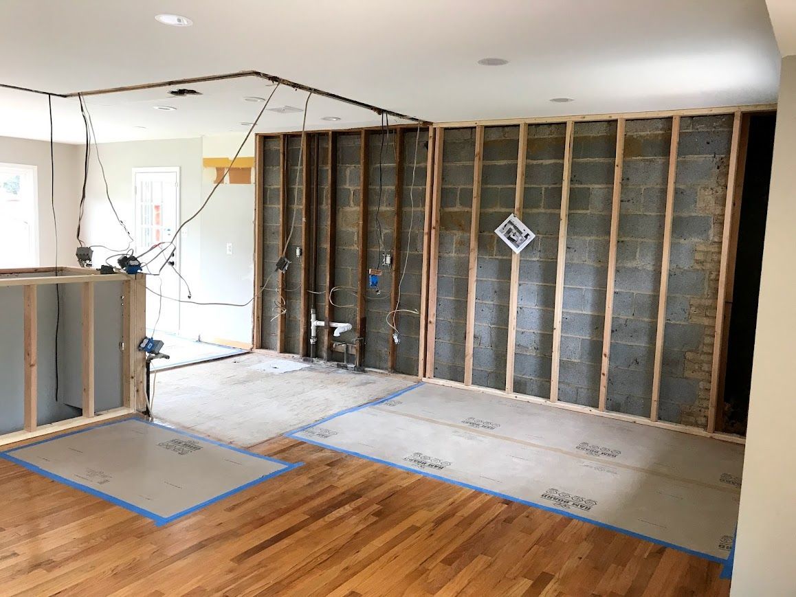 Interior room under renovation: exposed concrete block wall, wooden studs, wiring, and partially covered floor.