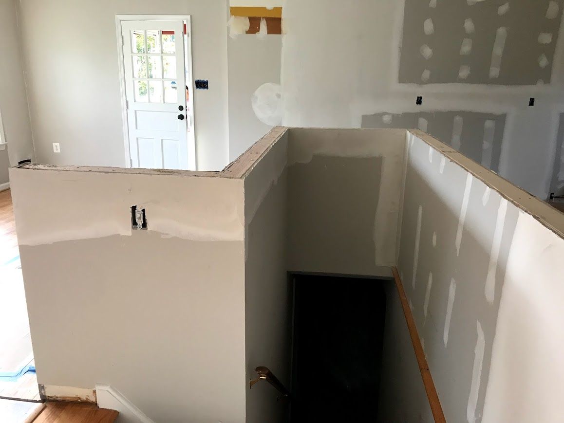 Interior of a house under construction. Drywall covers walls; staircase leads down to a dark doorway.