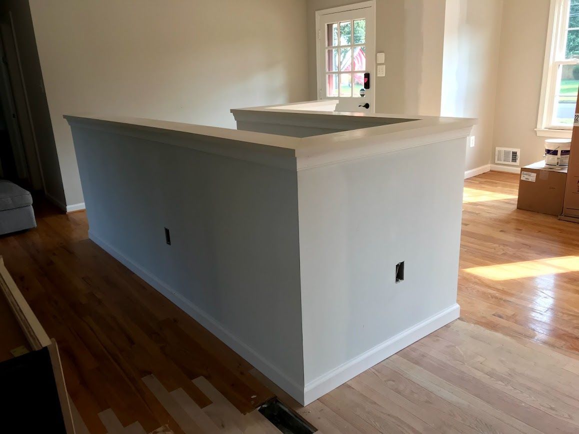 White wall with molding and electrical outlets, separating two rooms with hardwood floors.