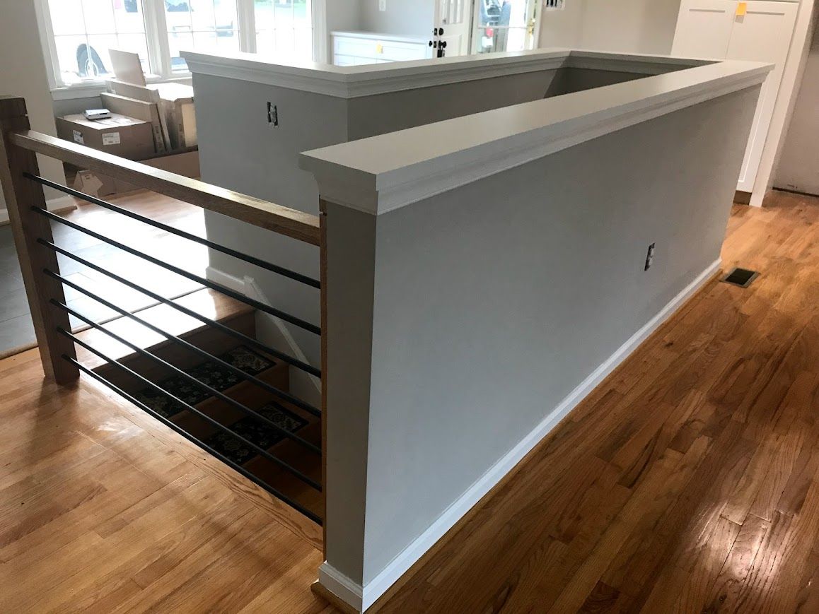 Interior view of a half-wall with a wooden handrail and black metal cables. Light gray walls, wood floors.