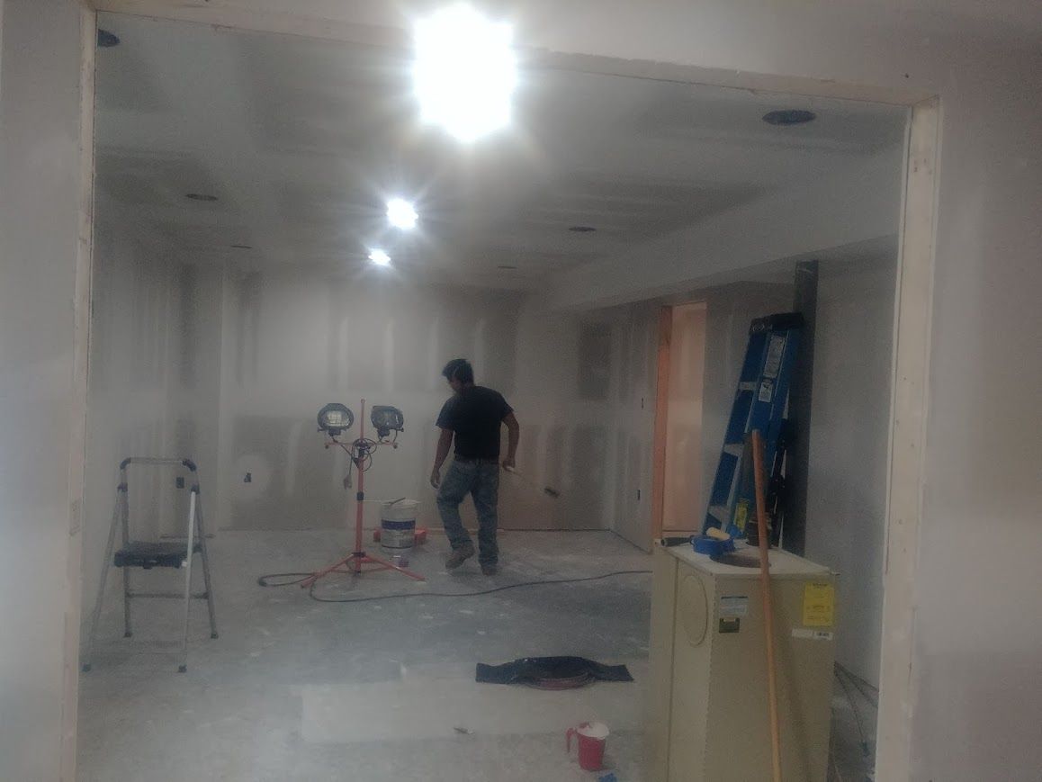 Room undergoing renovation, drywall visible. A person stands in the center with a work light.  Ladder and tools are present.
