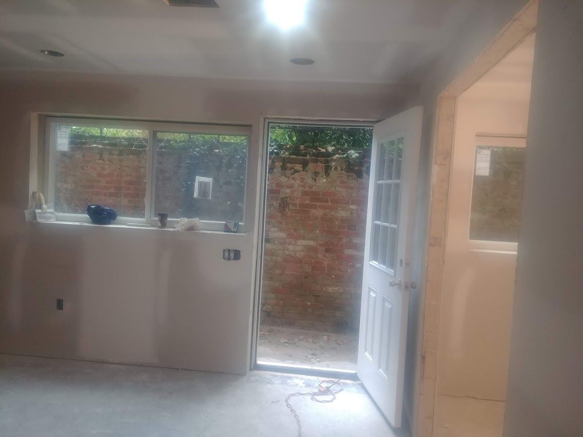 Interior view of a room under construction with a door and window. Drywall and brick are visible.