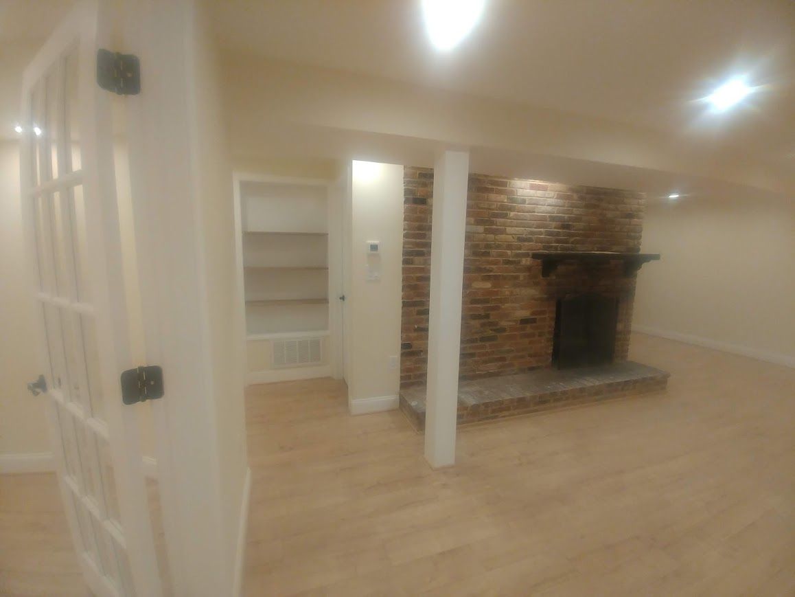 Basement room with fireplace, built-in shelving, and light wood flooring. White walls and trim.