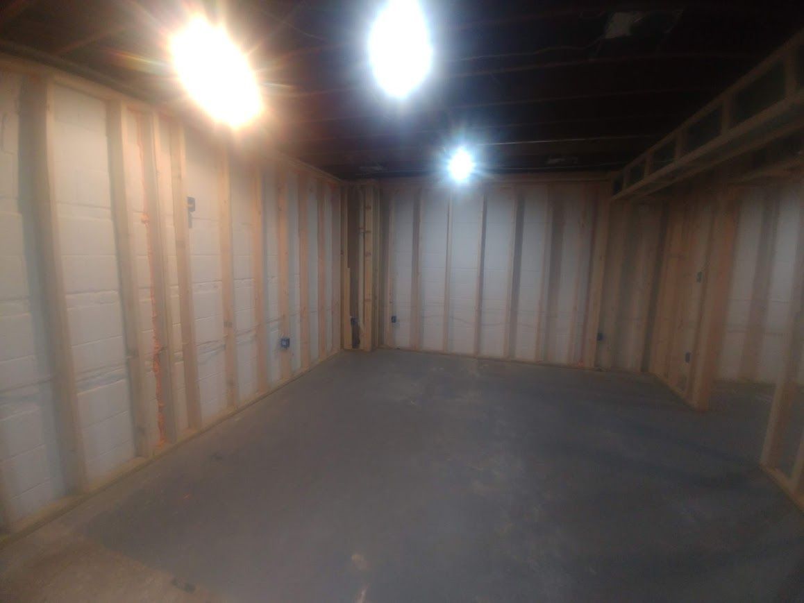 Unfinished basement interior with exposed studs, insulation, and concrete floor. Fluorescent lights shine from the ceiling.