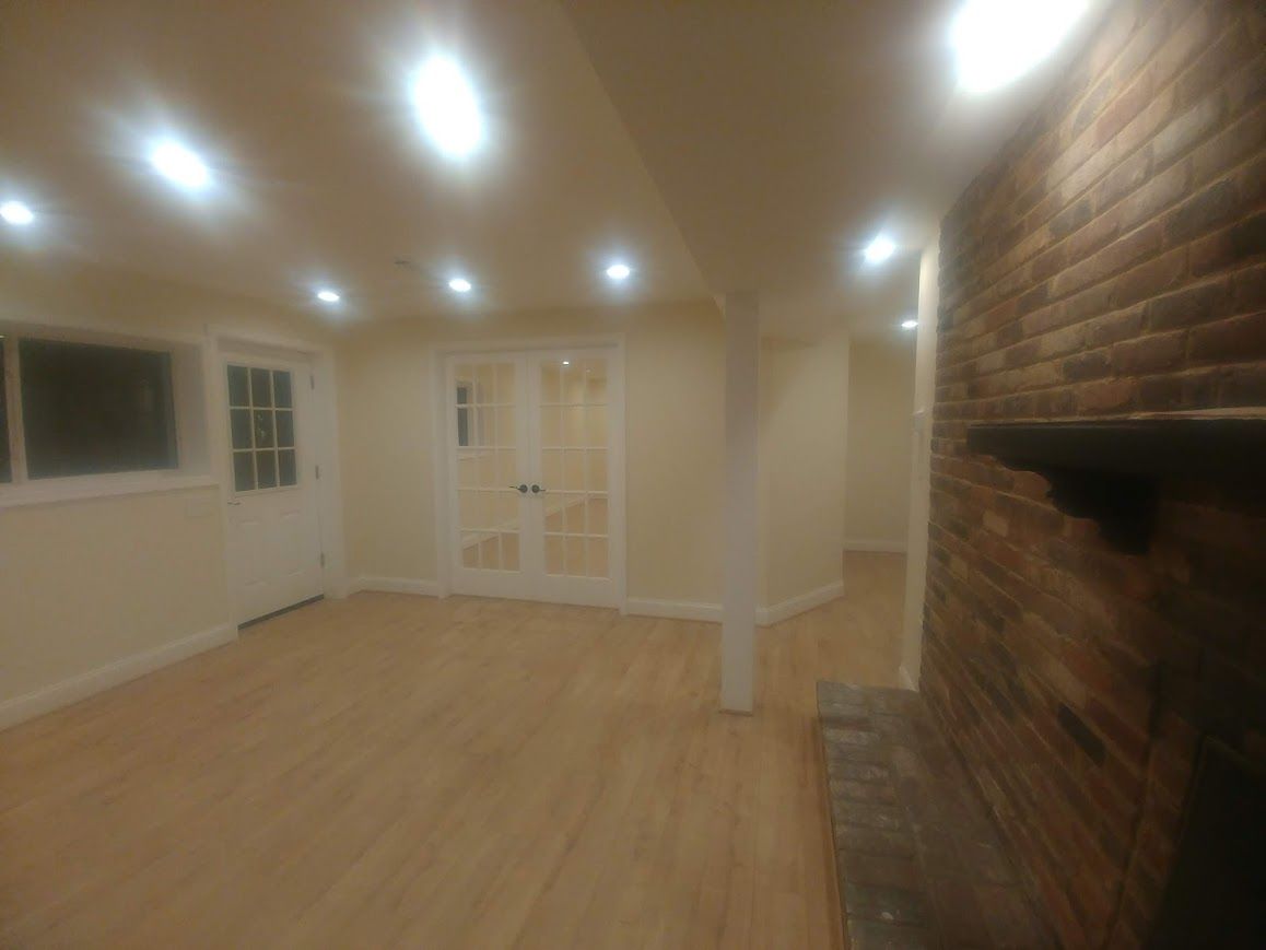 Finished basement with brick fireplace, light wood floors, recessed lighting, and doors.