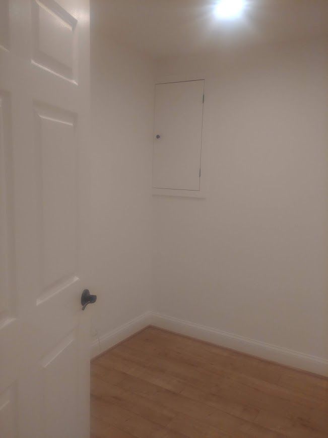 Small, empty room with wooden floor, white walls and a fuse box. A white door is on the left.