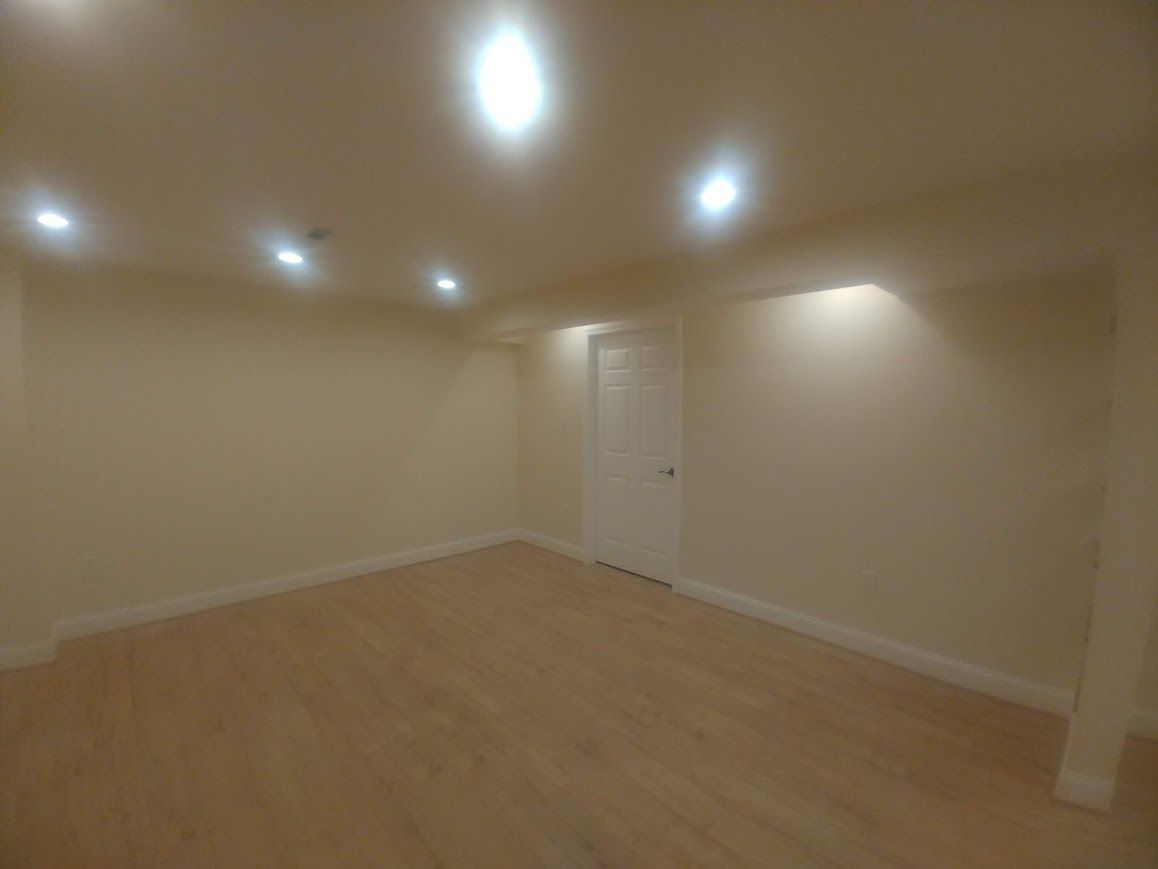Empty room with light-colored walls and wood flooring, door at the end, recessed ceiling lights.