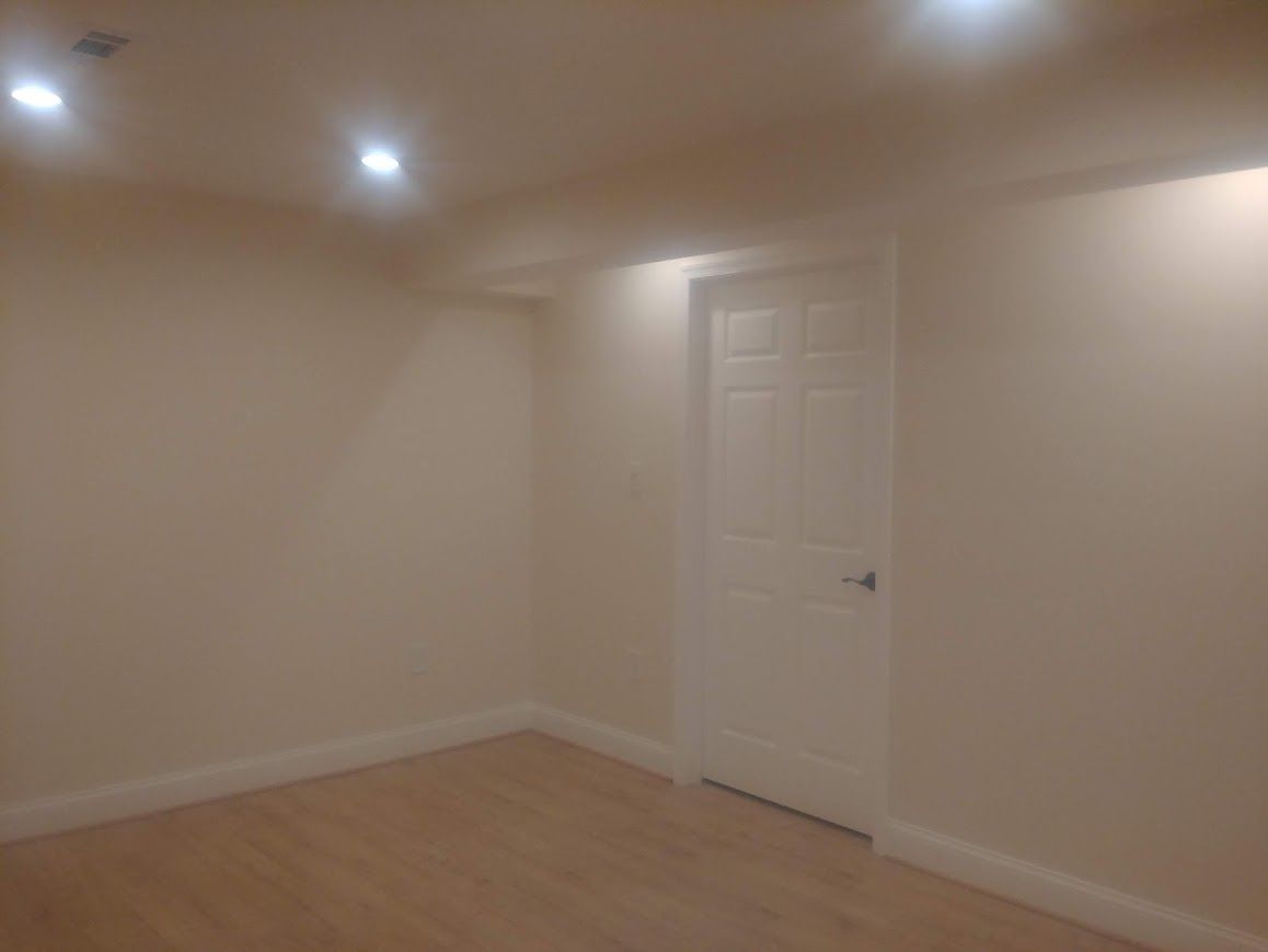 Empty room with white walls, recessed ceiling lights, white door, and light wood flooring.