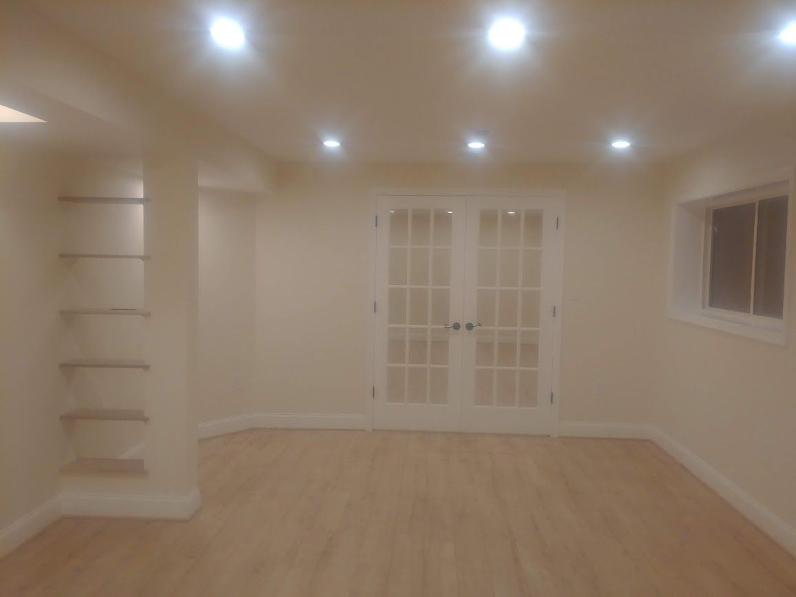 Empty, beige-colored room with recessed ceiling lights, French doors, wooden floors, and built-in shelving.