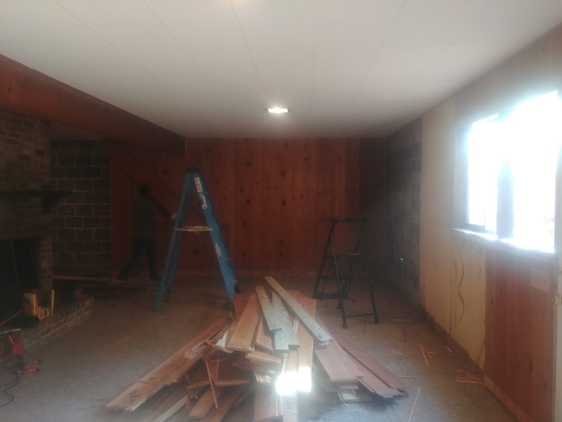 Room under renovation with wood paneling, boards on floor, ladder, and a window.