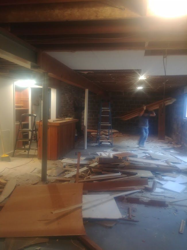 Man carrying lumber in a basement under construction; debris and exposed beams present.