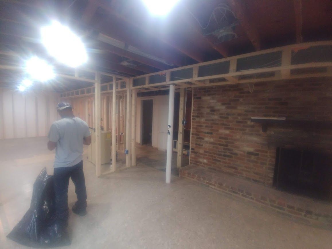 Basement under construction, with wooden frames, brick fireplace, and a person holding a trash bag.