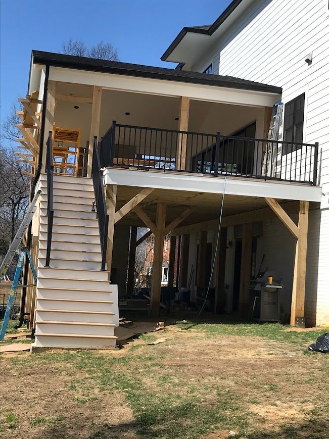 Two-story deck under construction with white siding, black railing, and wooden support beams on a sunny day.