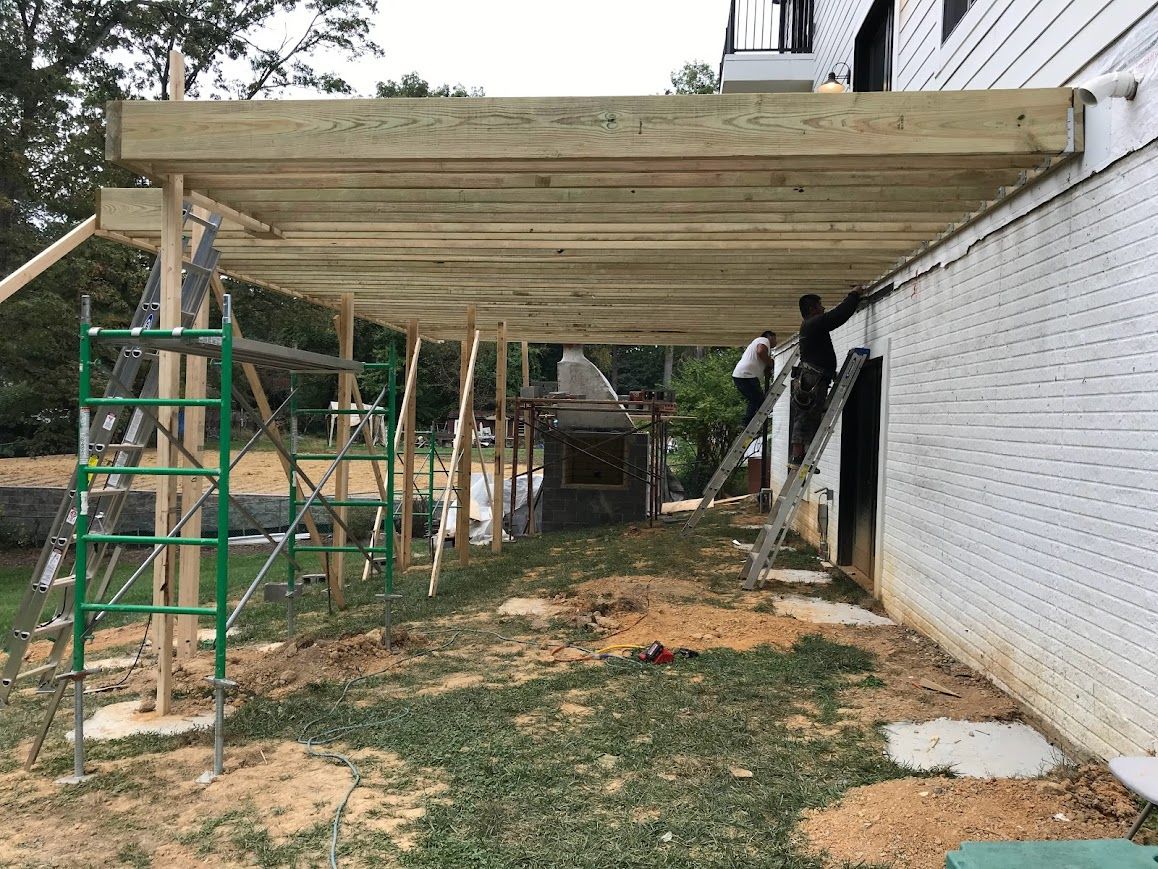 Construction of a wooden deck. Workers on scaffolding near a white brick wall and a concrete patio.