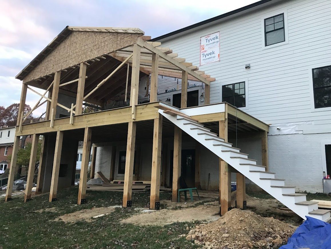 Construction of a raised wooden deck with stairs attached to a two-story white house; unfinished roof.