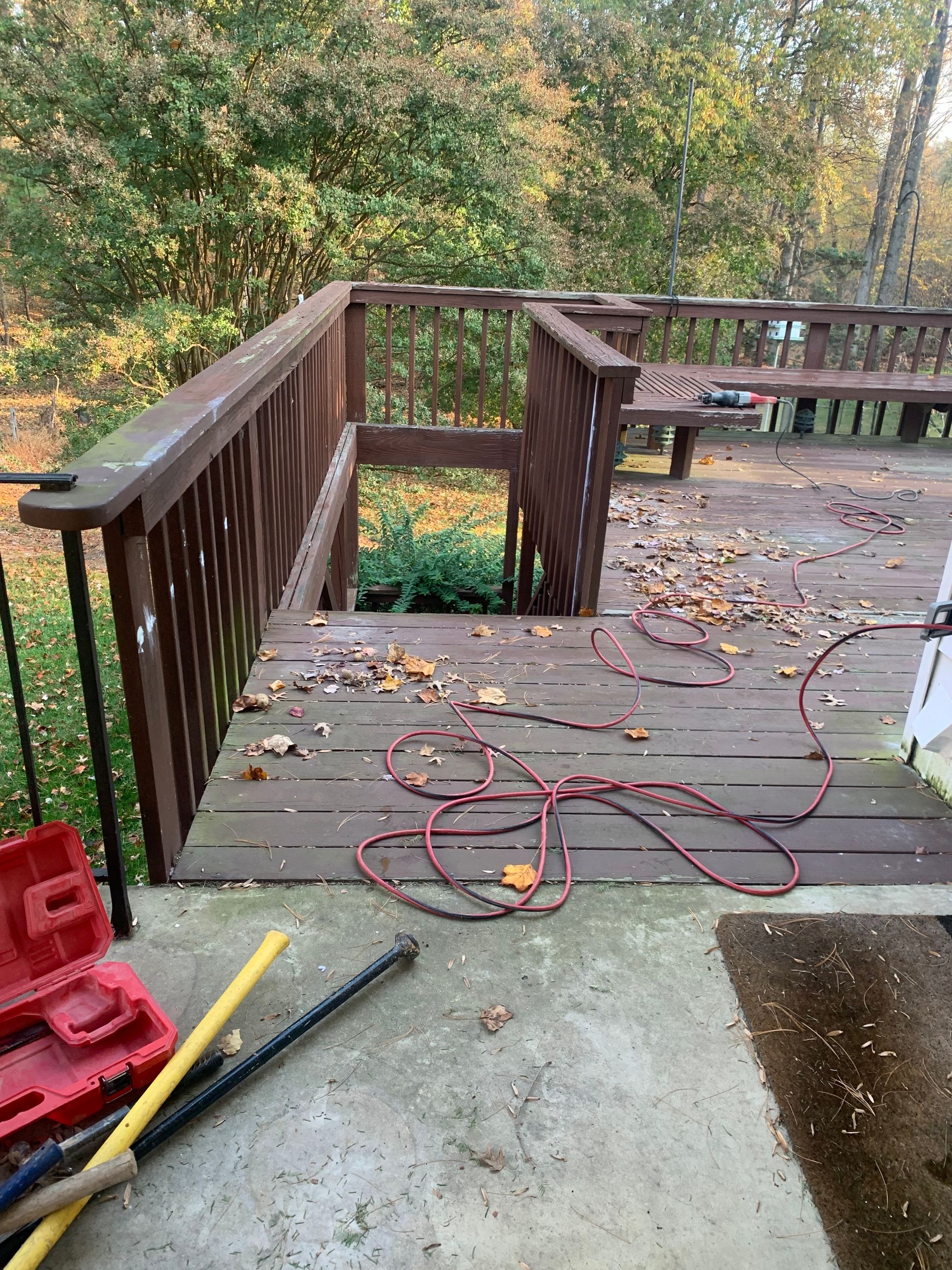 Deck with stairs, brown railings, fallen leaves, tools on concrete, power cord.