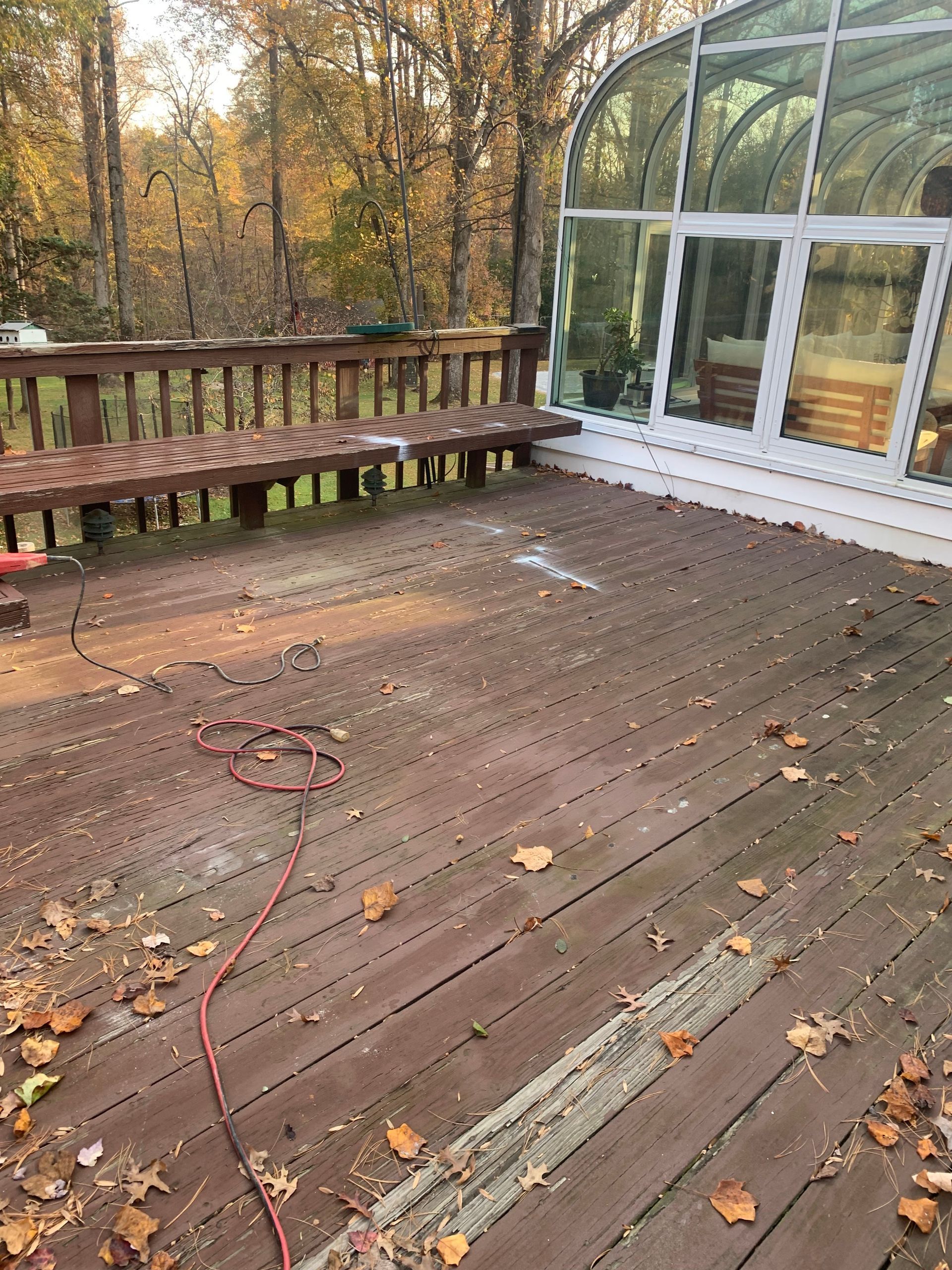 Brown wooden deck with bench, power cord, and a glass-enclosed structure. Fallen leaves and trees in the background.