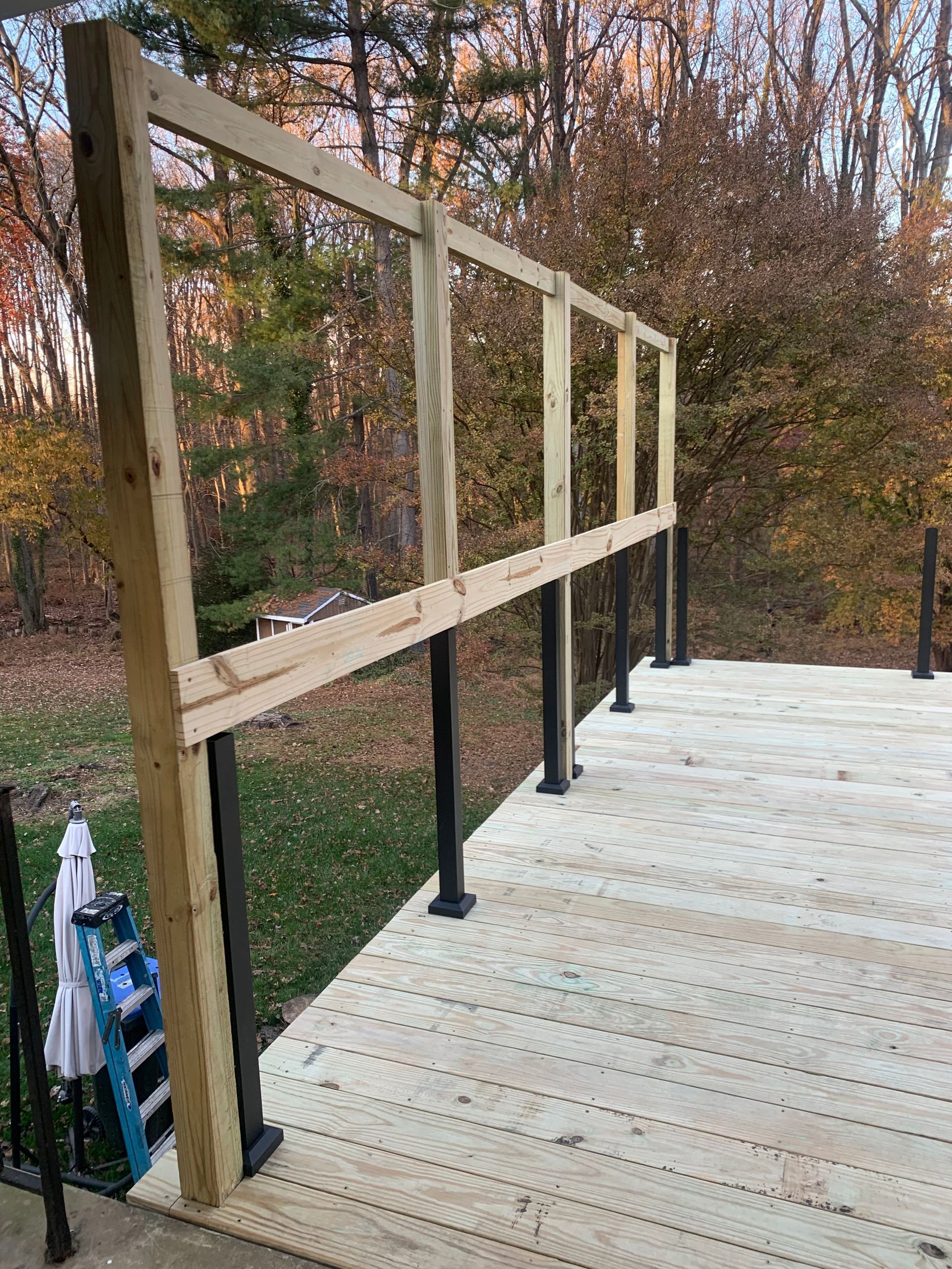 Wooden deck with black posts and unfinished wooden railing frame against a fall foliage backdrop.